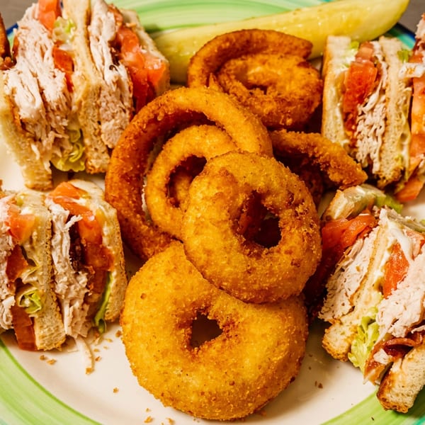 Four pieces of a club sandwich with onion rings and a pickle on a colorful plate.