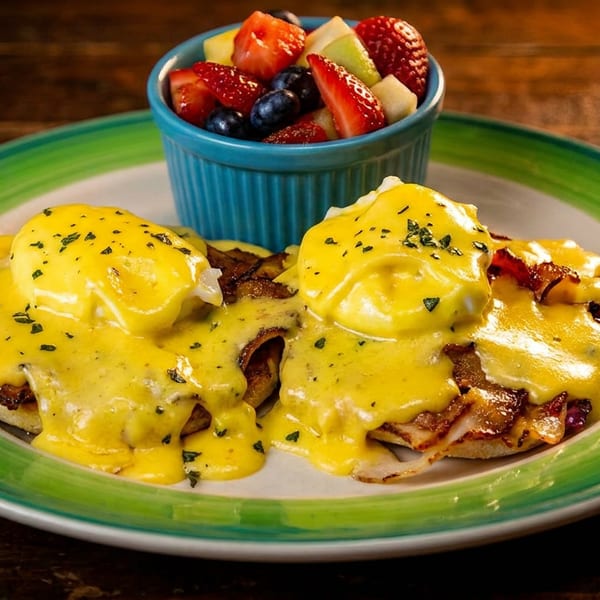 Two Eggs Benedict with hollandaise sauce and bacon, served with a side of fresh fruit.