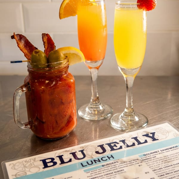 A Bloody Mary, two mimosas, and a Blu Jelly lunch menu on a stainless steel counter.