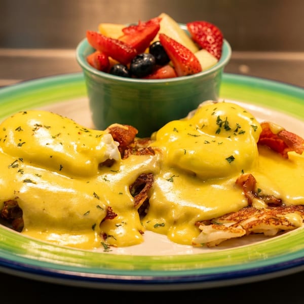 Two Eggs Benedict on crispy hash browns with hollandaise sauce and a side of fresh fruit.